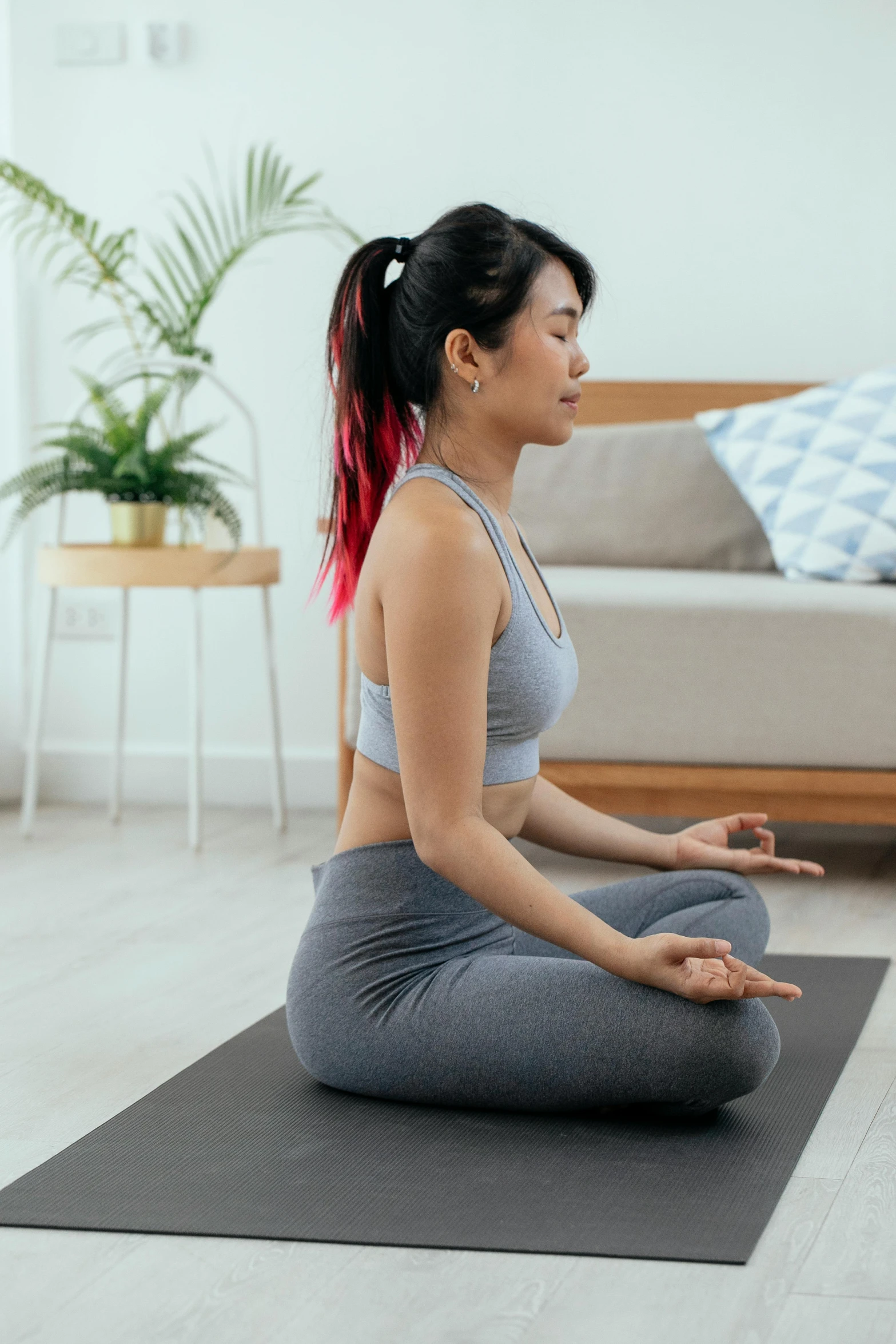 a woman sitting on a yoga mat in a living room, meditation pose, with a spine crown, asian features, wearing fitness gear