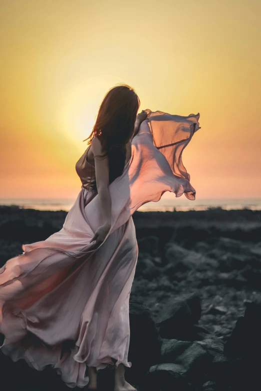a woman standing on top of a rocky beach, pexels contest winner, romanticism, flowing dress, pink sunset, windblown dark hair, ((sunset))