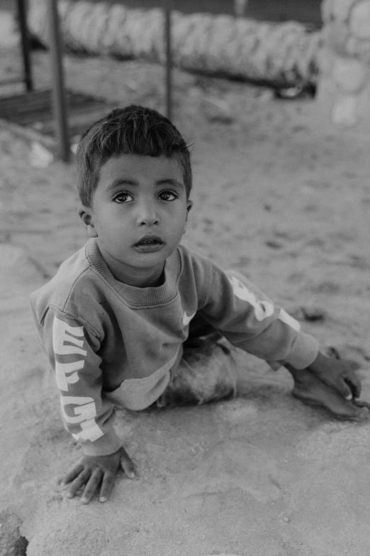 a black and white photo of a young boy, by Ahmed Yacoubi, sitting on the ground, portrait of a small character, on the sand, eyes looking at the camera