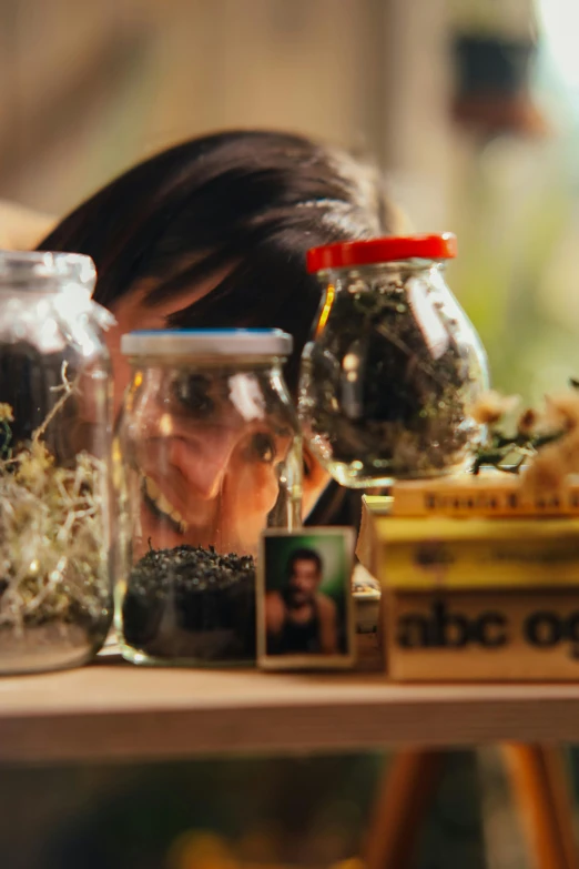 a woman looking through a glass jar filled with plants, still frame from a movie, lots of jars and boxes of herbs, thc, profile image