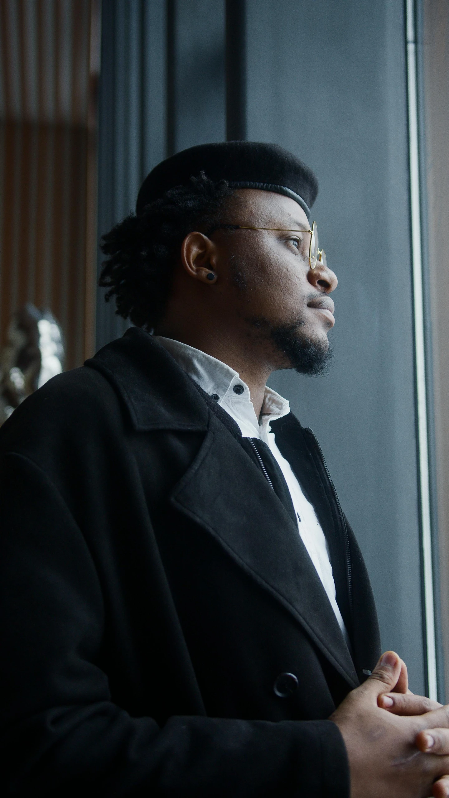 a man in a black coat looking out a window, an album cover, inspired by Edmond Aman-Jean, pexels, wakanda, man with glasses, wearing a beret, proud looking away