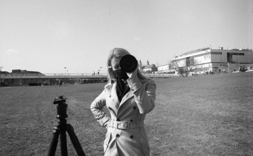 a black and white photo of a woman taking a picture, inspired by Henri Cartier-Bresson, on a racetrack, a blond, wearing trenchcoat, 1970s cinema camera