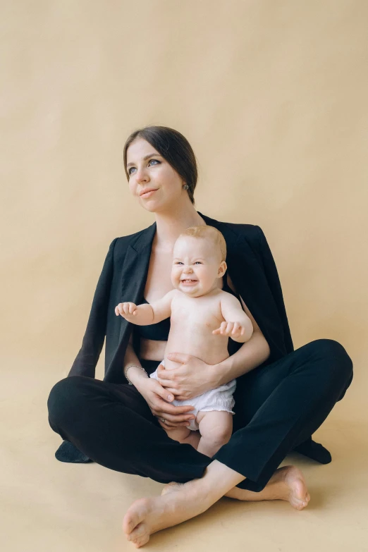 a woman sitting on the floor holding a baby, by Julia Pishtar, confident pose, solid background, pete davidson, portrait featured on unsplash