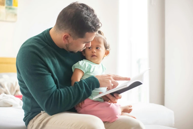 a man sitting on a bed reading a book to a baby, by Dan Content, pexels, renaissance, caring fatherly wide forehead, tiny girl looking on, holding notebook, comforting