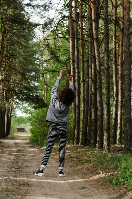 a woman reaching up in the air to catch a frisbee, by Grytė Pintukaitė, pexels contest winner, land art, tall trees, wearing a hoodie and sweatpants, handcrafted, standing in road
