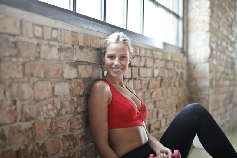 a woman in a red top and black leggings sitting against a brick wall, by Nina Hamnett, unsplash, dingy gym, bralette, smiling, manuka