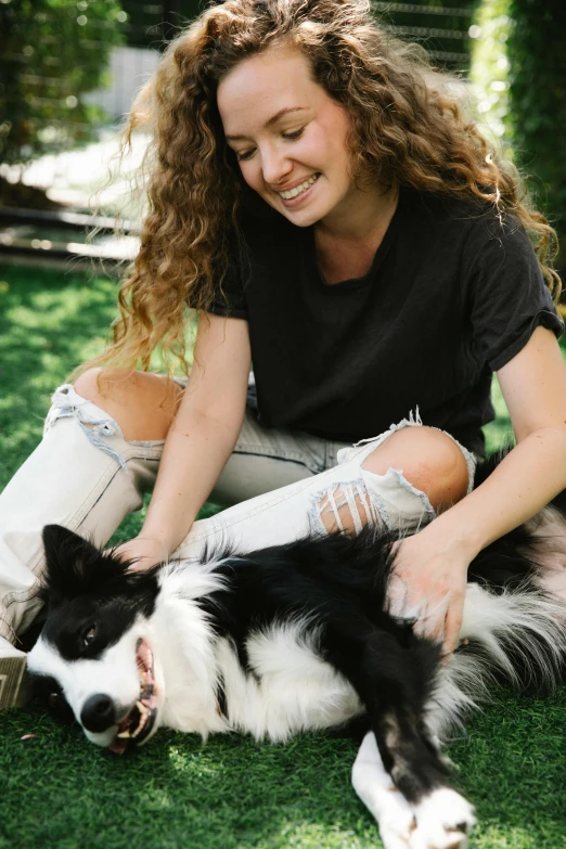 a woman sitting on the grass petting a black and white dog, jeans and t shirt, aussie, profile image, smiling down from above