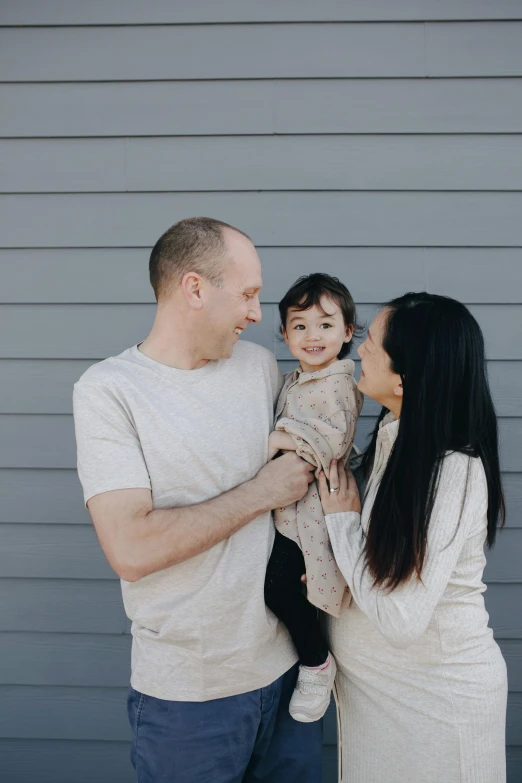 a man and woman holding a baby in front of a gray wall, pexels contest winner, standing outside a house, crisp smooth clean lines, sydney, white