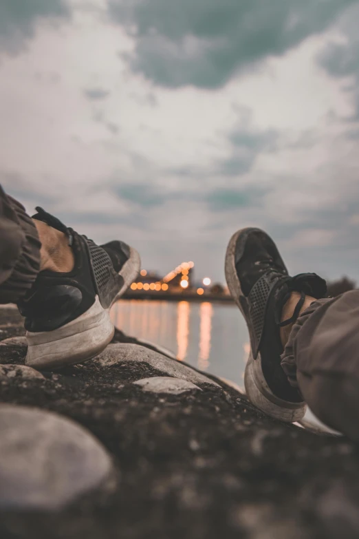 a person sitting on top of a rock next to a body of water, pexels contest winner, realism, sneaker shoes, warm summer nights, grey, festivals