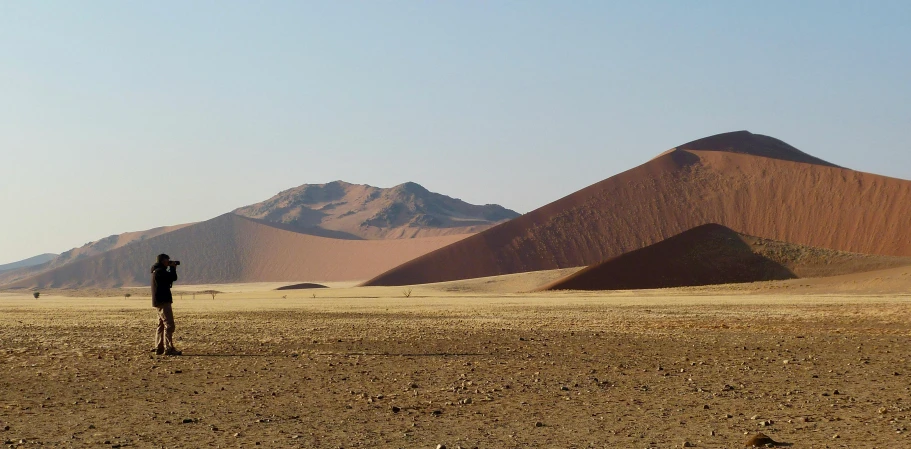 a man standing in the middle of a desert, by Peter Churcher, pexels contest winner, land art, giant crater in distance, panorama, landscape of africa, red peaks in the background