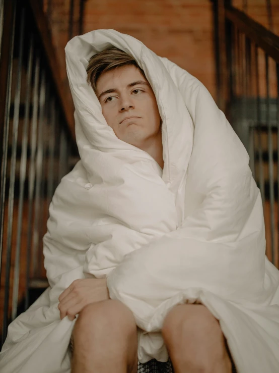 a man wrapped in a blanket sitting on a set of stairs, a colorized photo, inspired by Elsa Bleda, trending on pexels, joe keery, smirking deviously, lying in bed!!!! blanket, julian ope