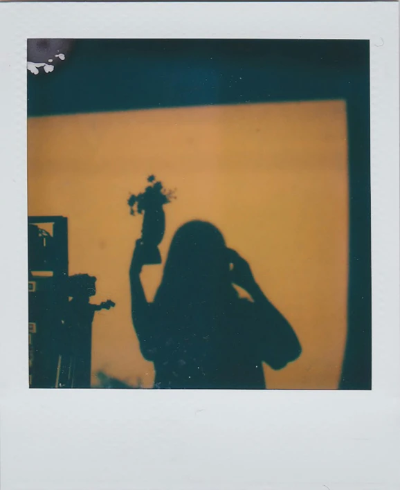 a woman holding a flower in front of a yellow wall, a polaroid photo, unsplash, silhouette :7, in a darkly lit laboratory room, ( ( photograph ) ), film cam