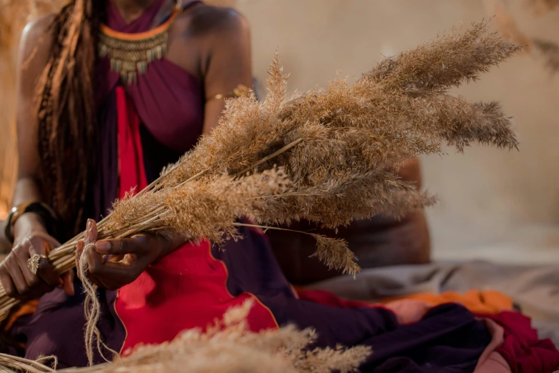 a woman sitting on a bed holding a bunch of dried grass, by Helen Stevenson, hurufiyya, blending, afro, an ancient, behind the scenes