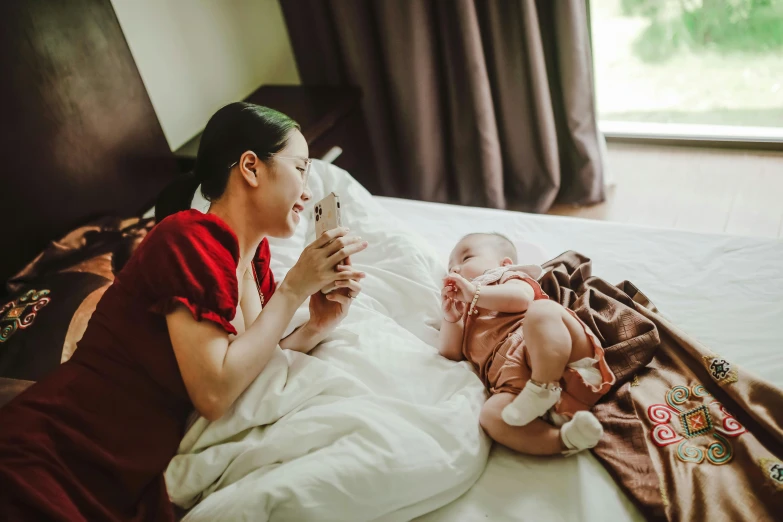 a woman laying on top of a bed next to a baby, by Julia Pishtar, pexels contest winner, happening, chinese heritage, looking at his phone, hotel room, avatar image