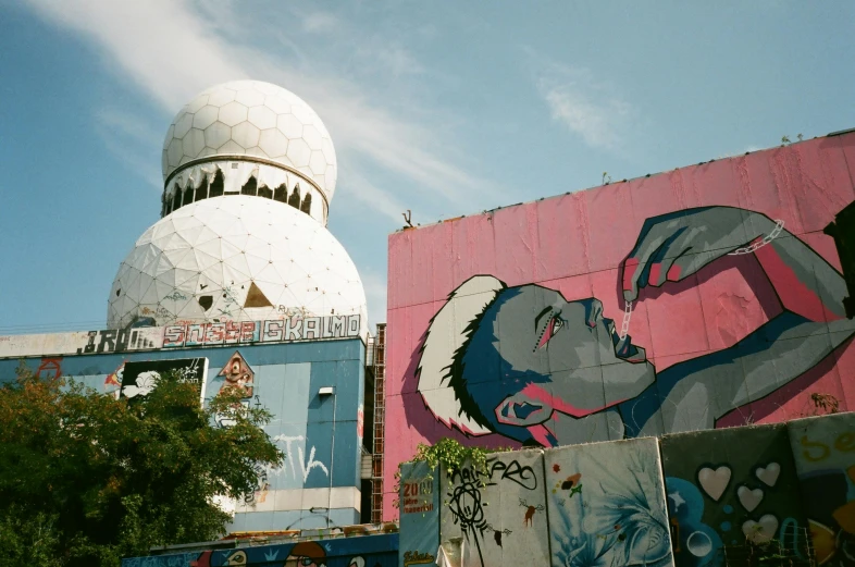 a mural of a man on the side of a building, inspired by Bhupen Khakhar, unsplash contest winner, berlin secession, big pink sphere high in the sky, 1980s photo, geodesic domes, towering above a small person