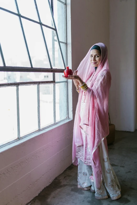 a woman standing in front of a window holding an apple, faridah malik, white and pink cloth, holding a bell, contemplative