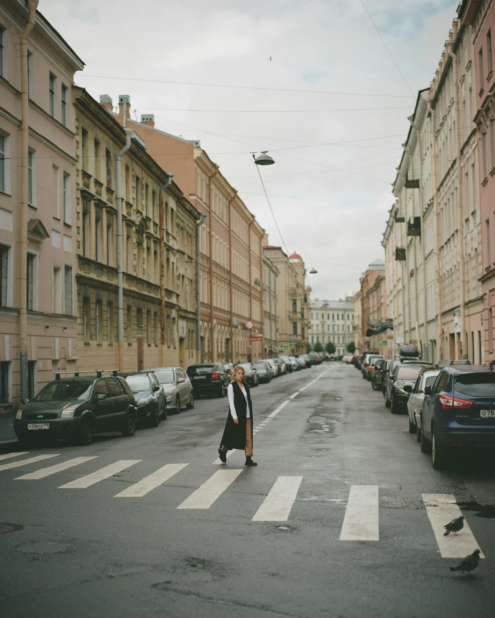 a woman walking across a street next to tall buildings, by Emma Andijewska, pexels contest winner, socialist realism, saint petersburg, non-binary, parking in the street, old town