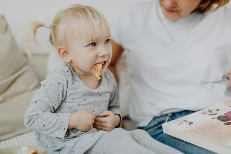 a woman is reading a book to a little girl, a picture, by Emma Andijewska, pexels contest winner, eating garlic bread, on grey background, sitting in his highchair, bite her lip