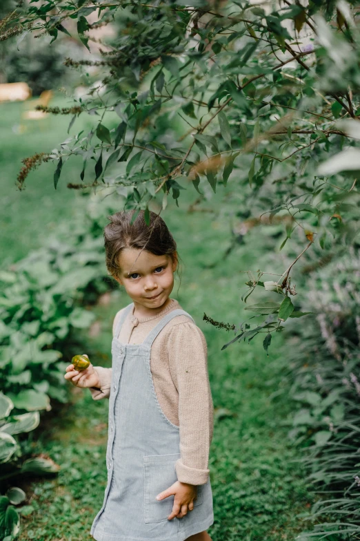 a little girl that is standing in the grass, inspired by Elsa Beskow, unsplash, garden with fruits on trees, wearing overalls, grey, candid portrait photo