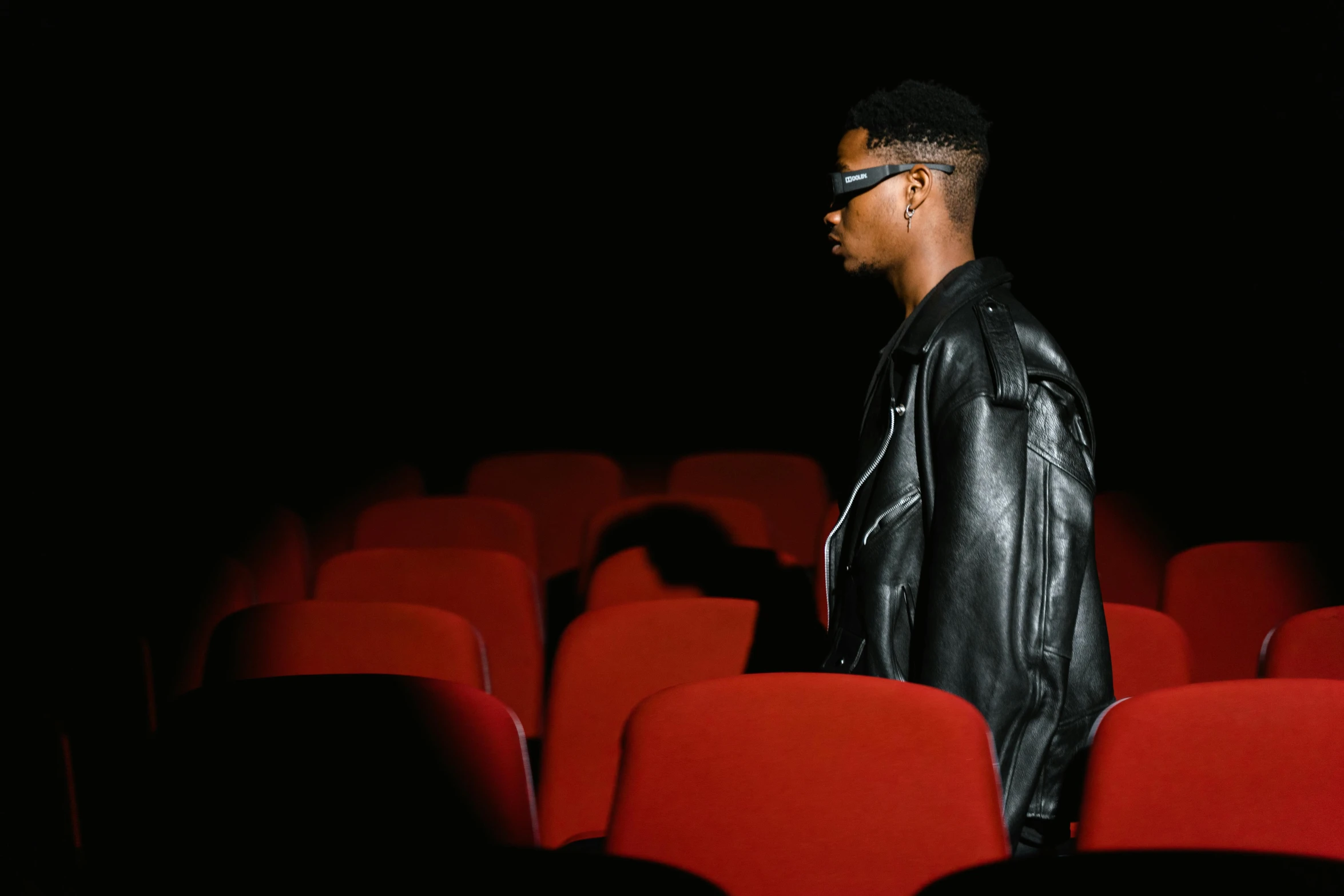 a man standing in front of a row of red chairs, an album cover, trending on pexels, visual art, portrait willow smith, sitting in a movie theater, dark shades, leather