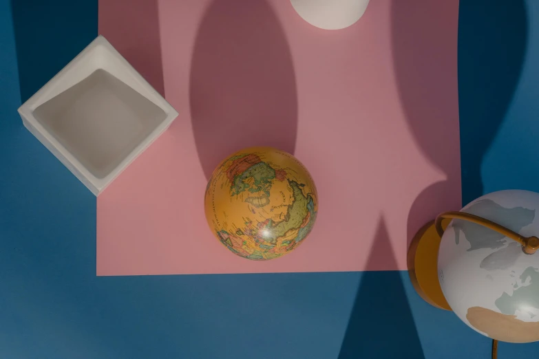 a globe sitting on top of a pink and blue surface, solid coloured shapes, sustainable materials, overhead sun, on a table