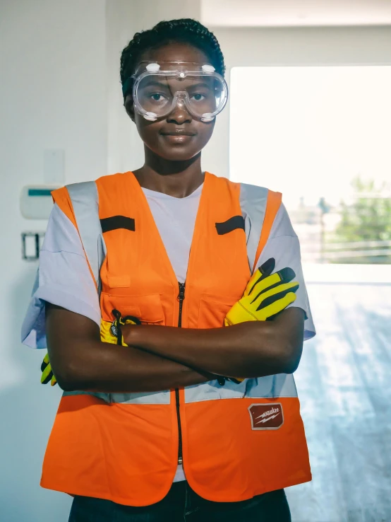 a woman in safety gear standing with her arms crossed, inspired by Afewerk Tekle, pexels contest winner, androgynous person, african american young woman, orange safety vest, wearing small round glasses