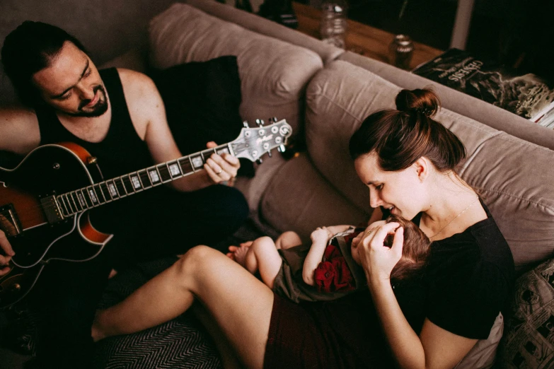 a man sitting on top of a couch next to a woman holding a baby, by Emma Andijewska, pexels contest winner, playing a gibson les paul guitar, evanescence, profile image, asleep