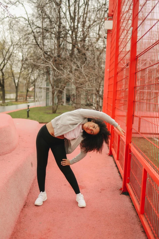 a woman standing in front of a red fence, doing splits and stretching, in russia, wearing a pastel pink hoodie, 2019 trending photo