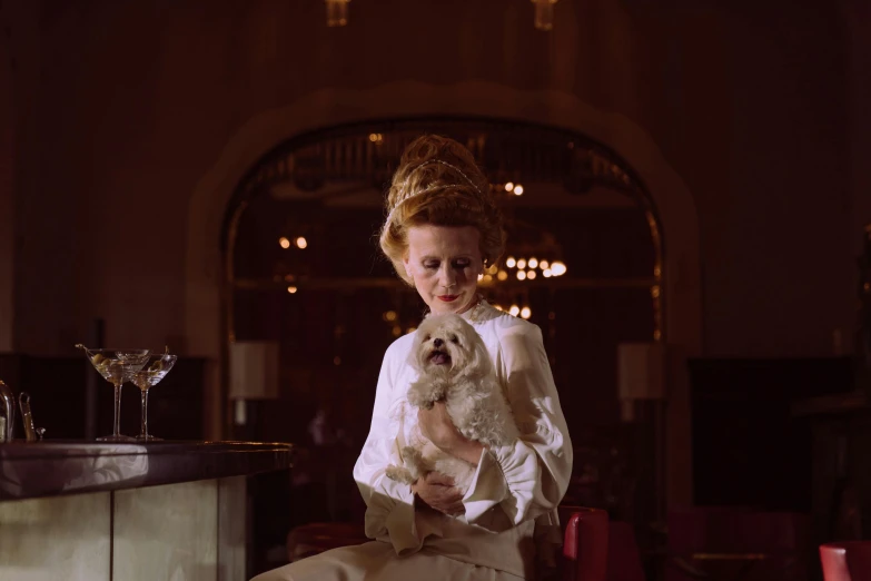 a woman sitting at a bar holding a dog, inspired by Félix Bódog Widder, trending on unsplash, renaissance, still from a wes anderson film, portrait tilda swinton, 1960s color photograph, futuristic ballroom. big eyes