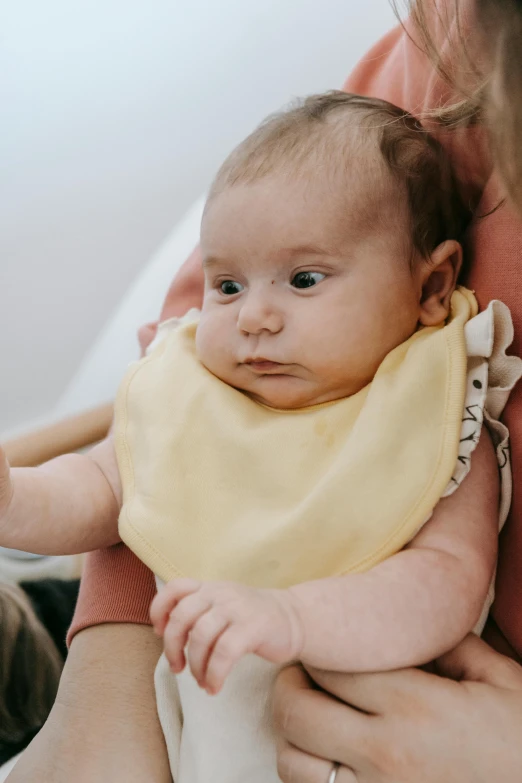 a woman holding a baby wearing a bib, pexels, with yellow cloths, head tilted down, sitting in chair, manuka