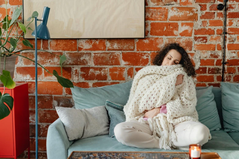 a woman sitting on a couch wrapped in a blanket, inspired by Elsa Bleda, pexels, happening, curly haired, bloated, wearing casual sweater, overflowing with diapers