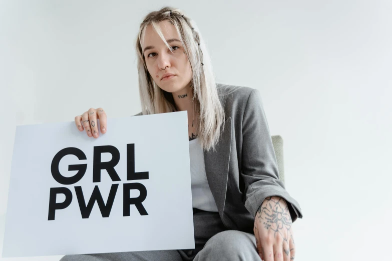 a woman holding a sign that says girl pwr, by Arabella Rankin, trending on pexels, gordon freeman as a woman, sitting down, grey, lily frank