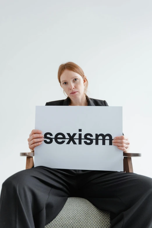 a woman sitting on a chair holding a sign that says sexism, trending on pexels, excessivism, on a gray background, with pale skin, profile image, scowling