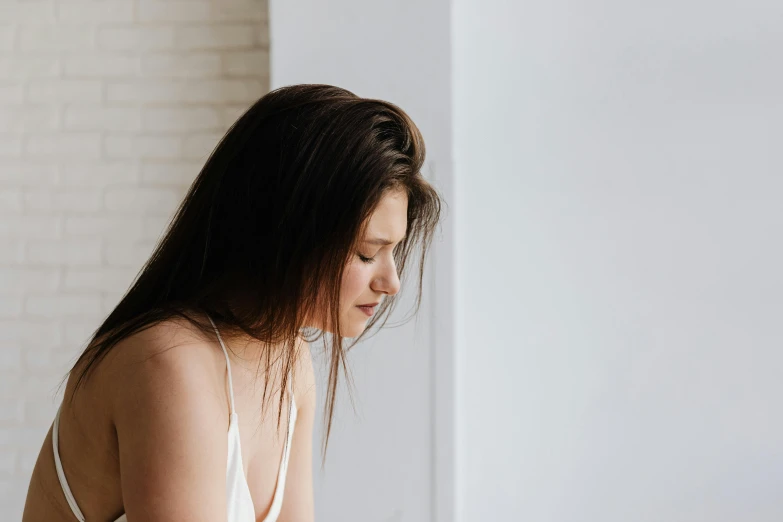 a woman in a white dress sitting on a bed, trending on pexels, happening, head turned to the side, girl with dark brown hair, disappointed, background image