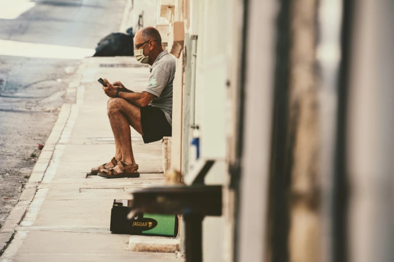 a man sitting on the side of a building looking at his cell phone, a photo, by Matt Stewart, pexels contest winner, hyperrealism, old cmputers on the sidewalk, wearing facemask and sunglasses, trying to read, brown