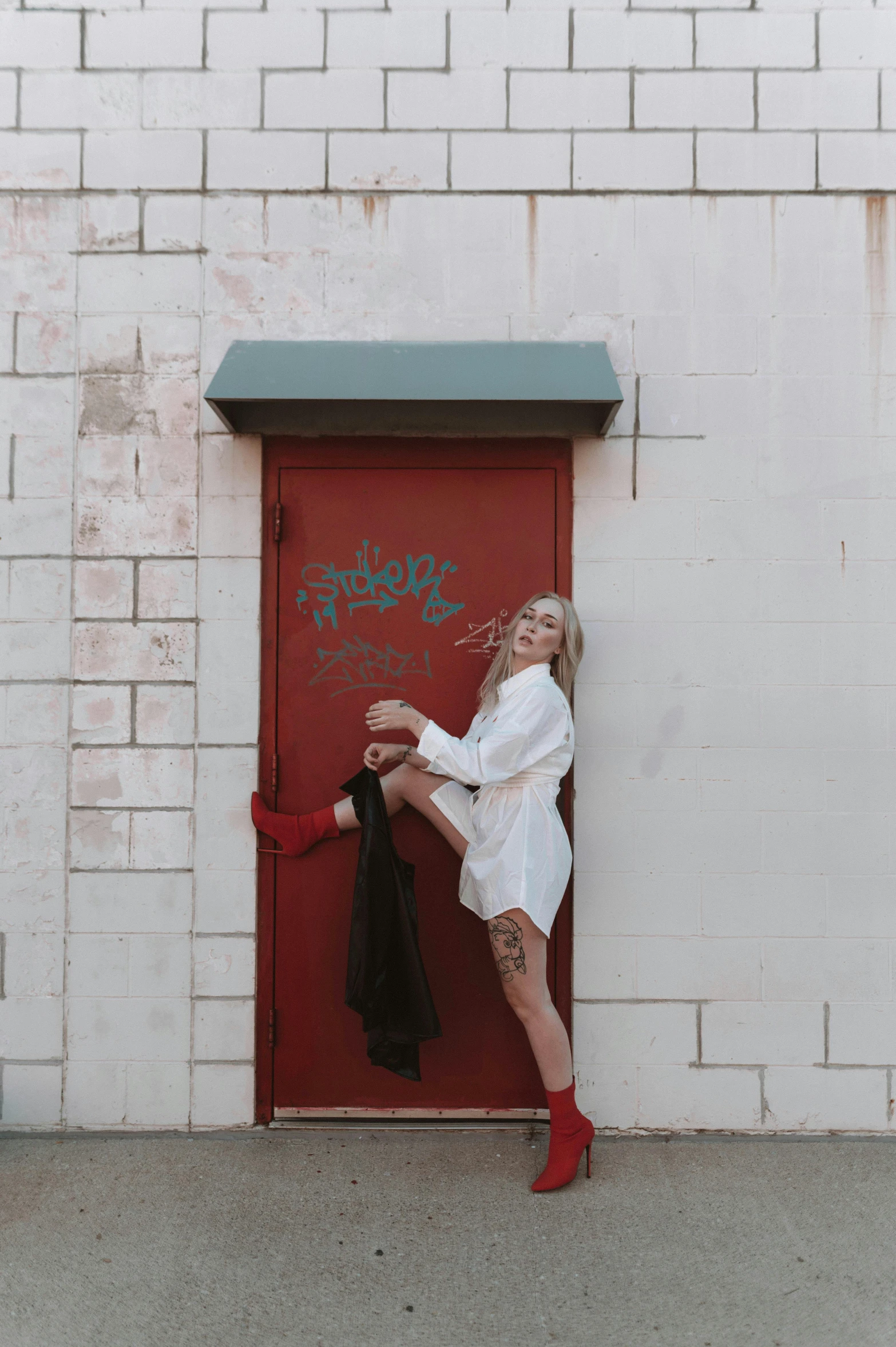 a woman in a white dress leaning against a red door, a photo, by Jessie Alexandra Dick, pexels contest winner, graffiti, she wears boots, platinum blonde, white backdrop, playful pose