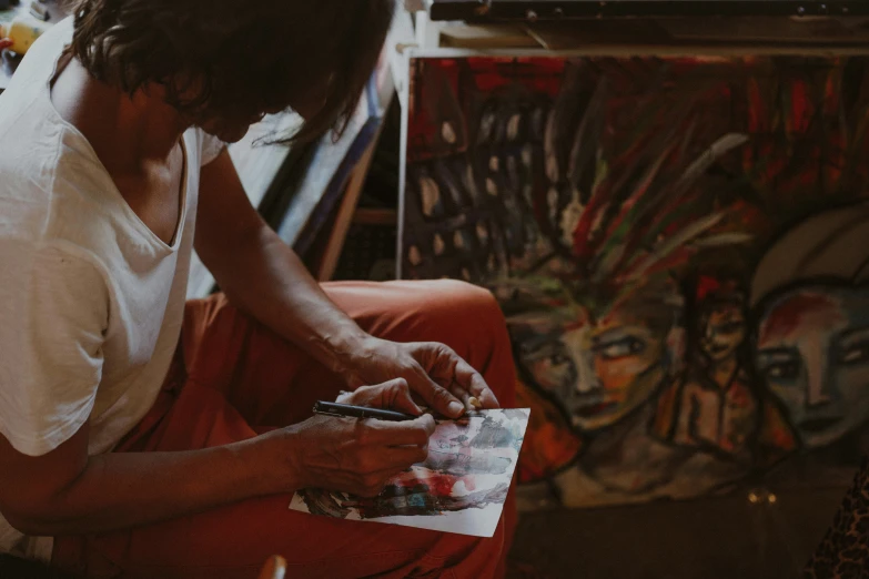 a woman sitting on a chair in front of a painting, pexels contest winner, process art, drawing sketches on his notebook, graffiti painting, studious chiaroscuro, indigenous art