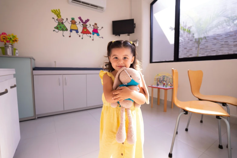 a little girl in a yellow dress holding a teddy bear, a cartoon, inspired by Pacita Abad, pexels contest winner, doctors office, wide-shot, colour photograph, beige