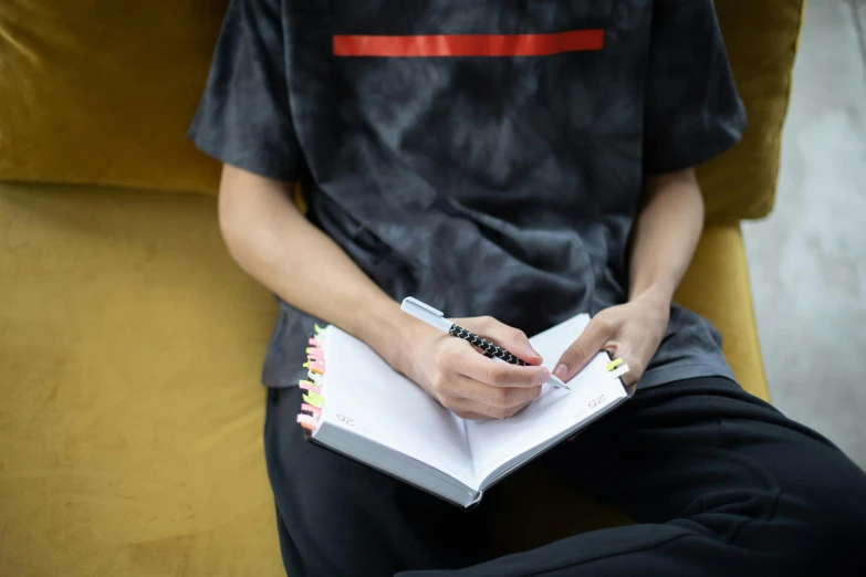 a man sitting on a yellow chair holding a notebook, a drawing, by Robbie Trevino, pexels contest winner, streetwear, close up half body shot, copic markers, academic clothing