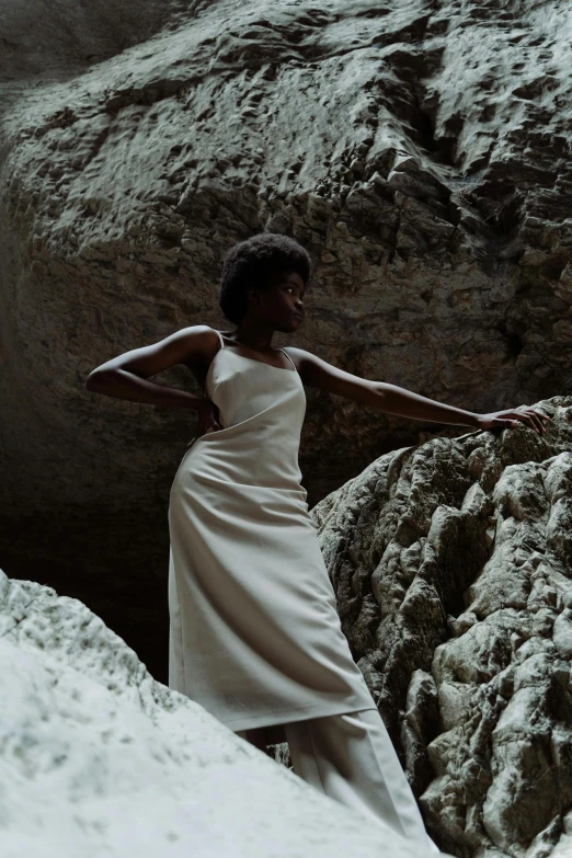 a woman in a white dress standing on a rock, inspired by Gabriel Ba, unsplash, afrofuturism, wearing an evening gown, muted brown, ignant, photo of a model