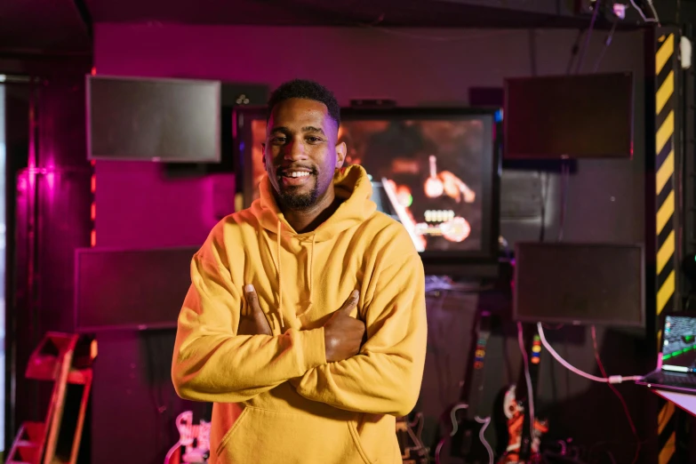 a man in a yellow hoodie standing with his arms crossed, by Stokely Webster, happening, in a nightclub, portrait image, mkbhd, he is wearing a brown sweater
