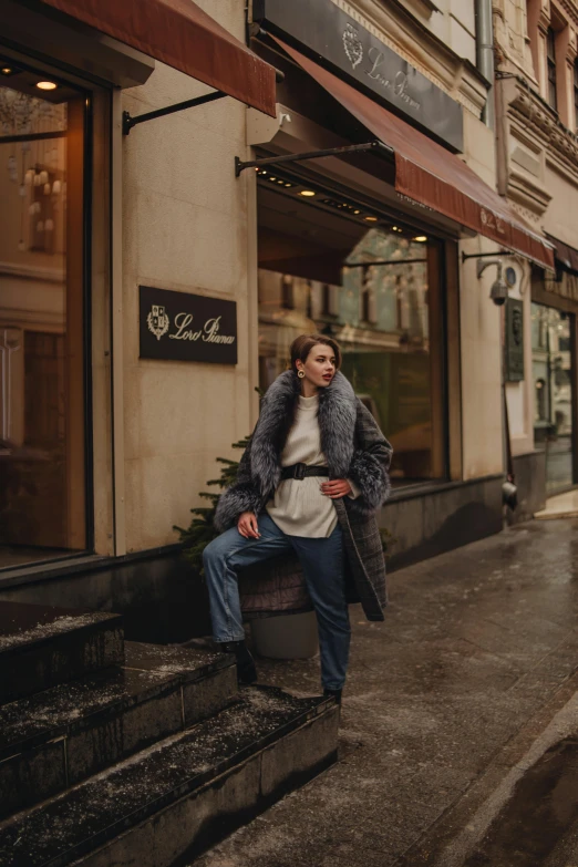 a woman sitting on a ledge in front of a building, inspired by Peter Lindbergh, trending on pexels, realism, fur jacket, exiting store, russian style, standing in street