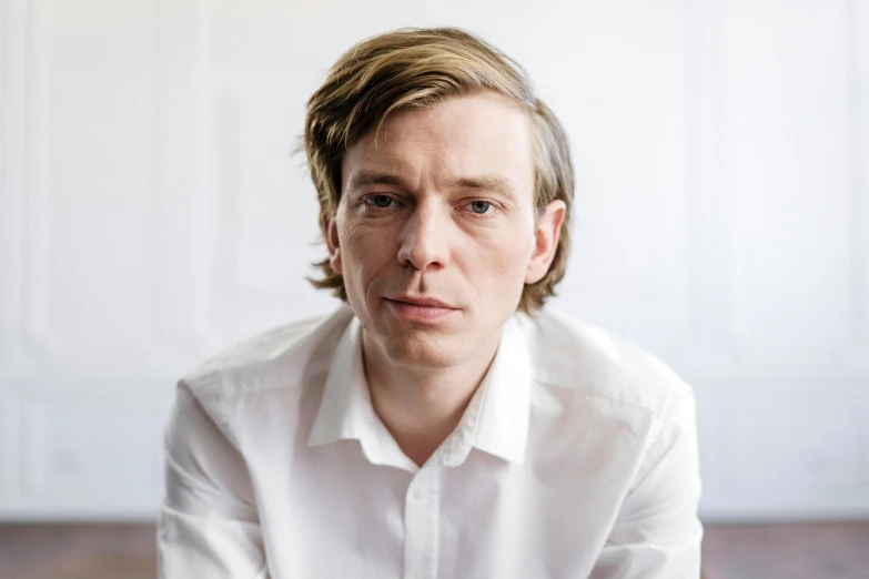 a man in a white shirt sitting on a wooden floor, by Alexander Kanoldt, unsplash, renaissance, looks like domhnall gleeson, alvar aalto, headshot photograph, julian ope