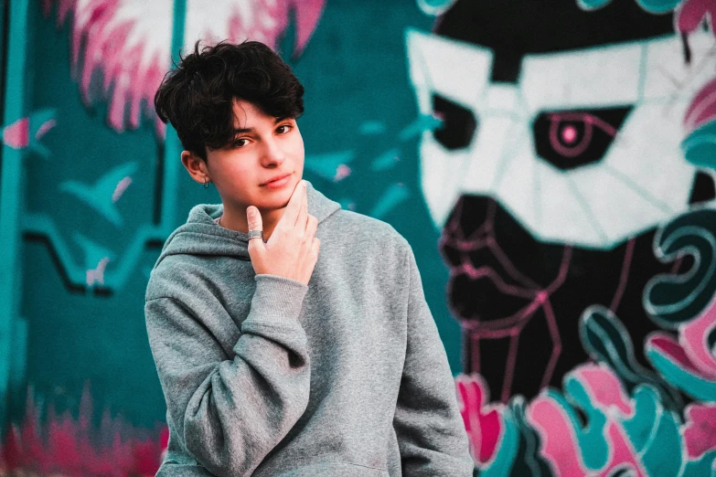 a young man standing in front of a graffiti covered wall, an album cover, inspired by John Luke, trending on pexels, graffiti, black haired girl wearing hoodie, cute boys, avatar image, ethan klein