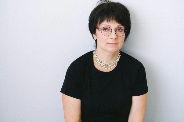 a woman sitting in front of a white wall, wearing black rimmed glasses, maria panfilova, with short hair, plain background