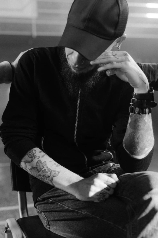 a man with a beard sitting in a chair, a black and white photo, inspired by Cam Sykes, pexels contest winner, photograph of a sleeve tattoo, baggy clothing and hat, large)}], devastated