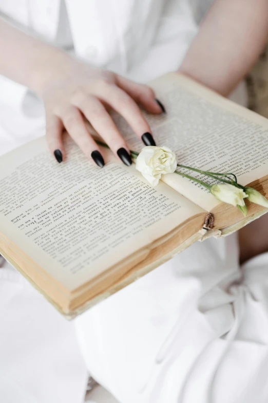 a woman sitting on a bed holding an open book, by Lucia Peka, unsplash, romanticism, white roses, sleek hands, gothic style, wedding