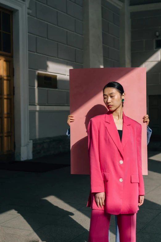 a woman in a pink suit standing in front of a building, an album cover, by Julia Pishtar, pexels contest winner, a portrait of issey miyake, chengyou liu, afternoon sun, egor letov