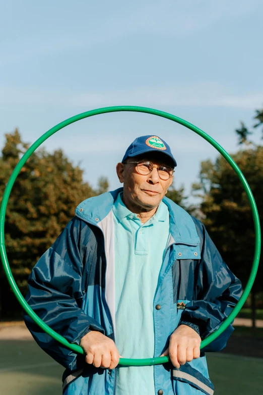 a man standing on a tennis court holding a hula hoop, he is about 8 0 years old, taejune kim, wearing green jacket, 2019 trending photo