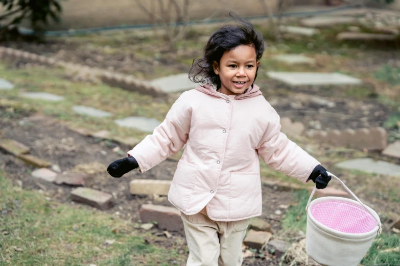 a little girl that is holding a bucket, inspired by Myles Birket Foster, pexels contest winner, wearing a pastel pink hoodie, walking at the garden, model wears a puffer jacket, ameera al-taweel